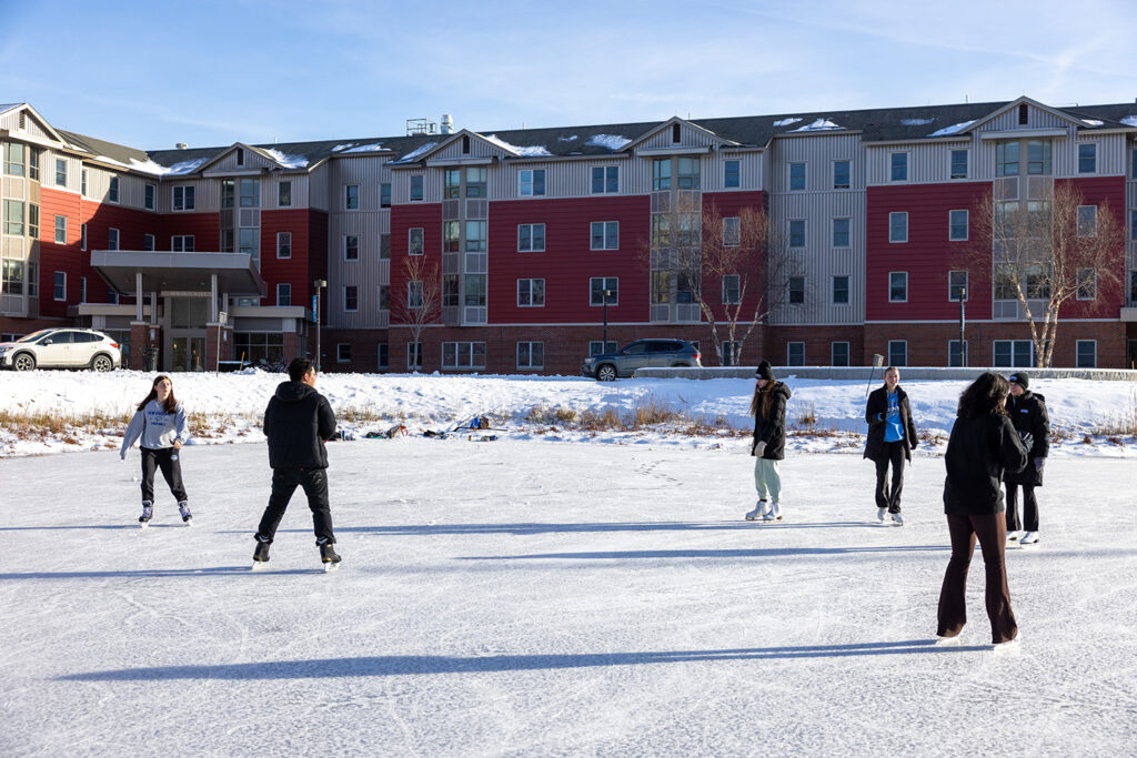 Students ice skate on outdoor campus rink in front of modern residence halls with red and gray siding on a sunny winter day.