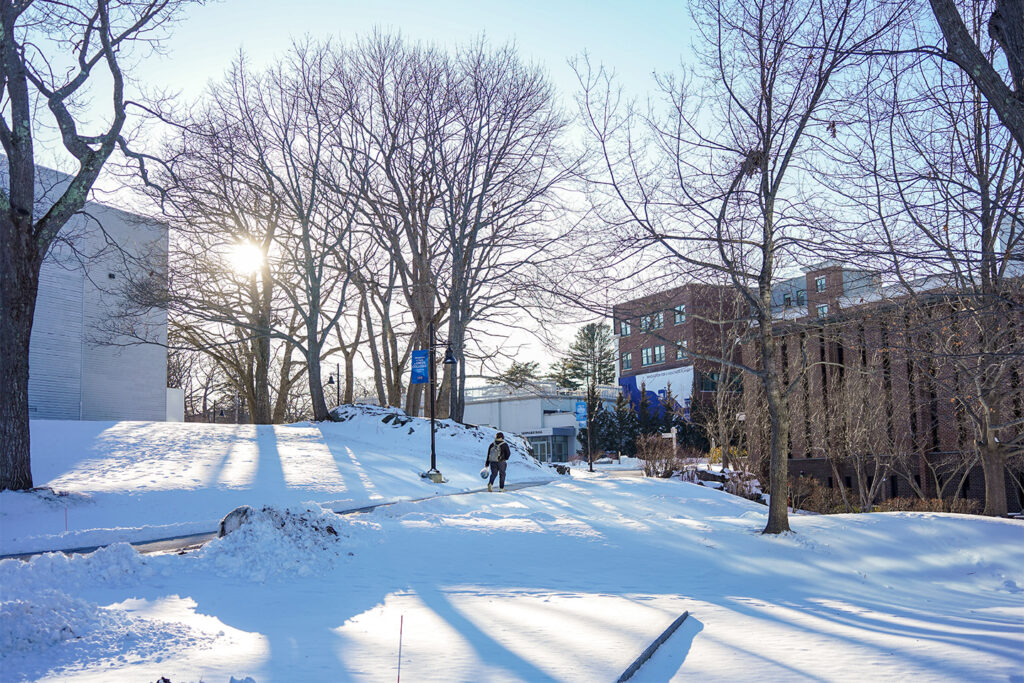 Student walks through snow-covered campus pathway on a bright winter morning, with bare trees casting long shadows and campus buildings visible in the background.