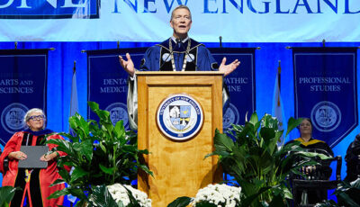 President Herbert speaks from a podium on stage with other university professionals sitting on either side