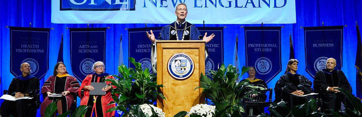 President Herbert speaks from a podium on stage with other university professionals sitting on either side