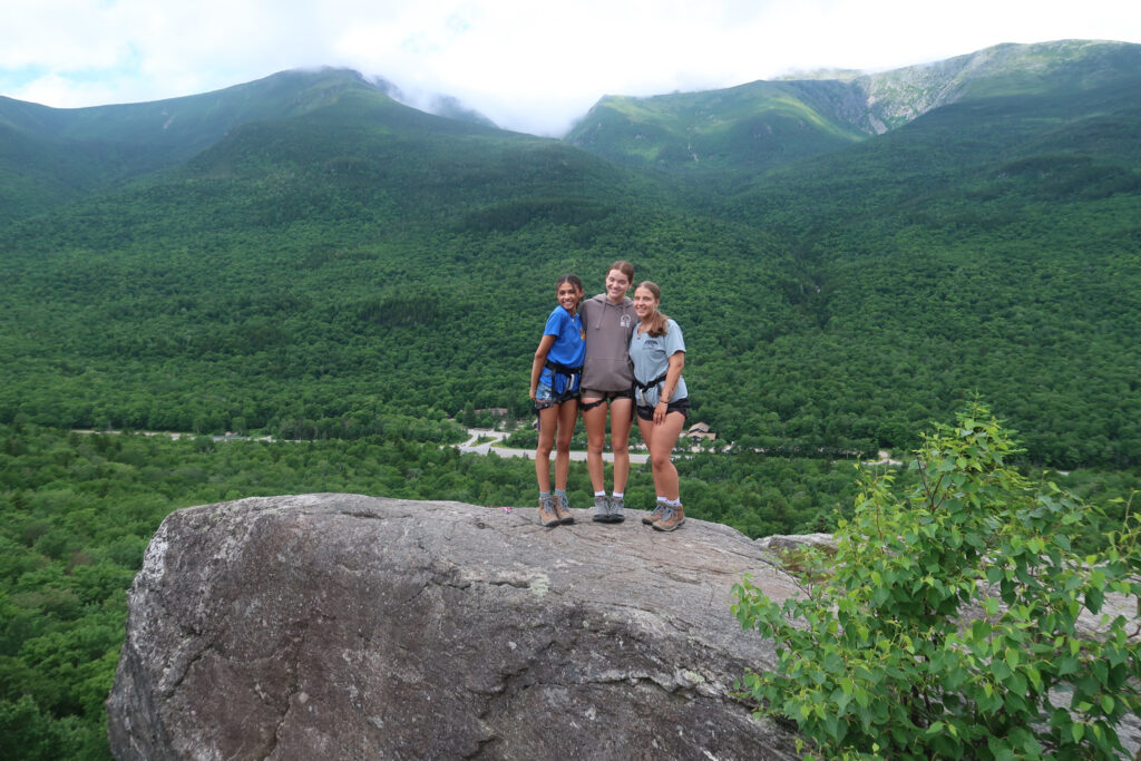 Three students standing on a rock in front of a forest and mountain backdrop