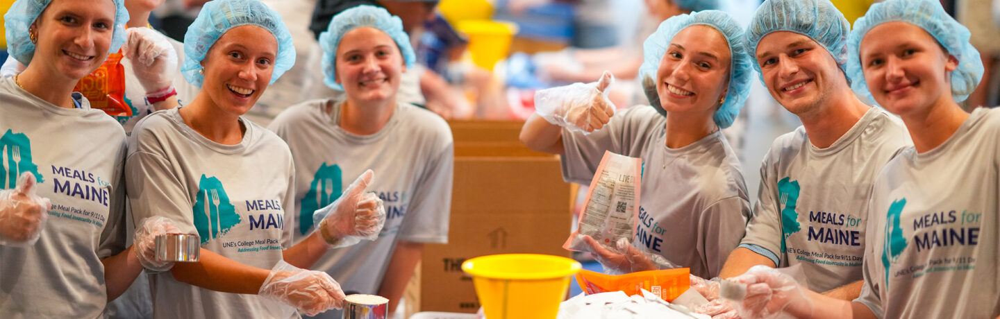 Group of volunteers wearing hairnets and Meals for Maine t-shirts package food at a community service event.