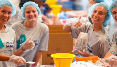 Group of volunteers wearing hairnets and Meals for Maine t-shirts package food at a community service event.