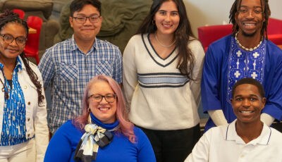 Group of six students and staff pose together in a campus lounge area with modern furniture and casual seating arrangements.