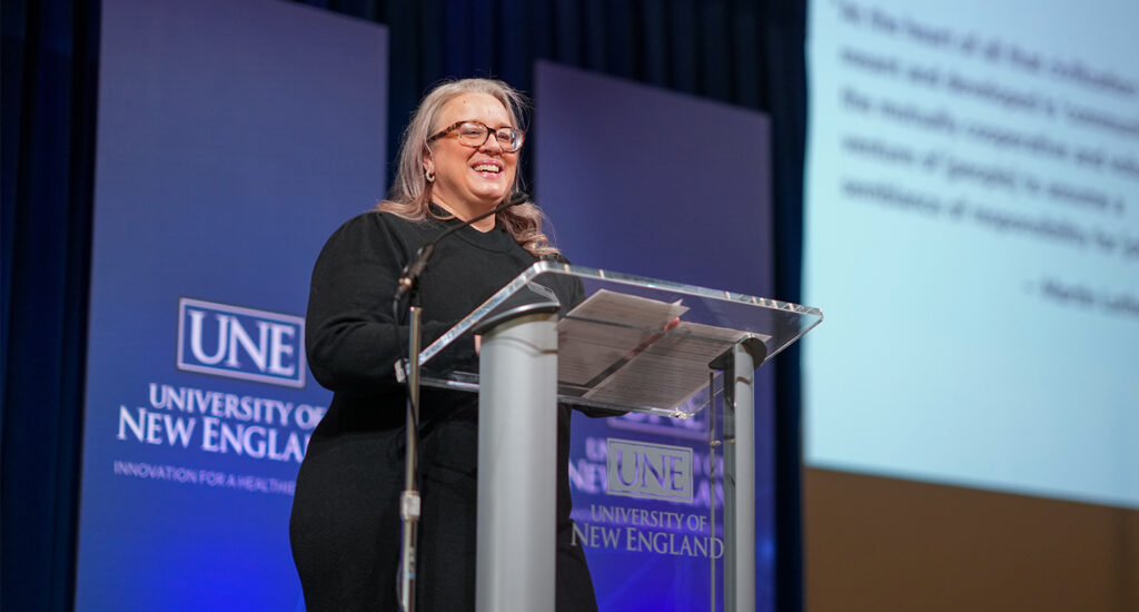Shannon Zlotkowski delivers remarks from a podium at an MLK Celebration event at the University of New England, with branded backdrop and presentation slides visible on screen.