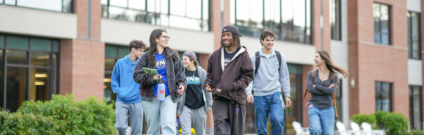 Group of six students walk together across campus in casual clothing and UNE apparel, with modern brick academic buildings and landscaping in the background.