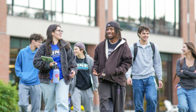 Group of six students walk together across campus in casual clothing and UNE apparel, with modern brick academic buildings and landscaping in the background.
