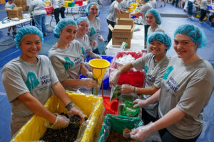 Group of volunteers wearing hairnets and Meals for Maine t-shirts package food into colored bins during community service event.
