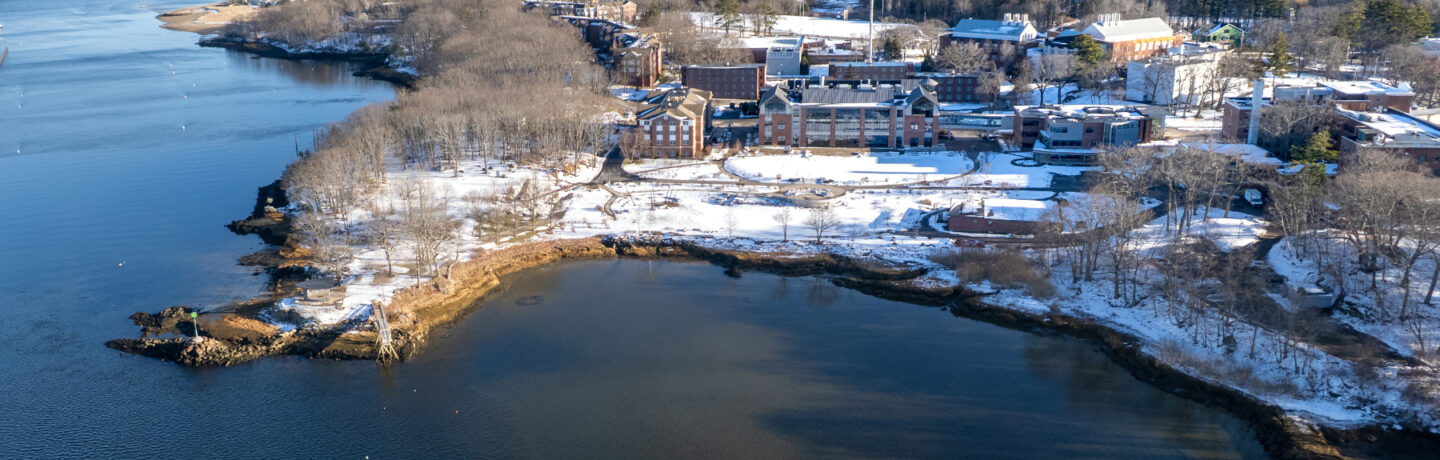 Aerial view of snow-covered Biddeford Campus waterfront with historic brick buildings, bare winter trees, and blue harbor waters.