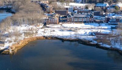 Aerial view of snow-covered Biddeford Campus waterfront with historic brick buildings, bare winter trees, and blue harbor waters.