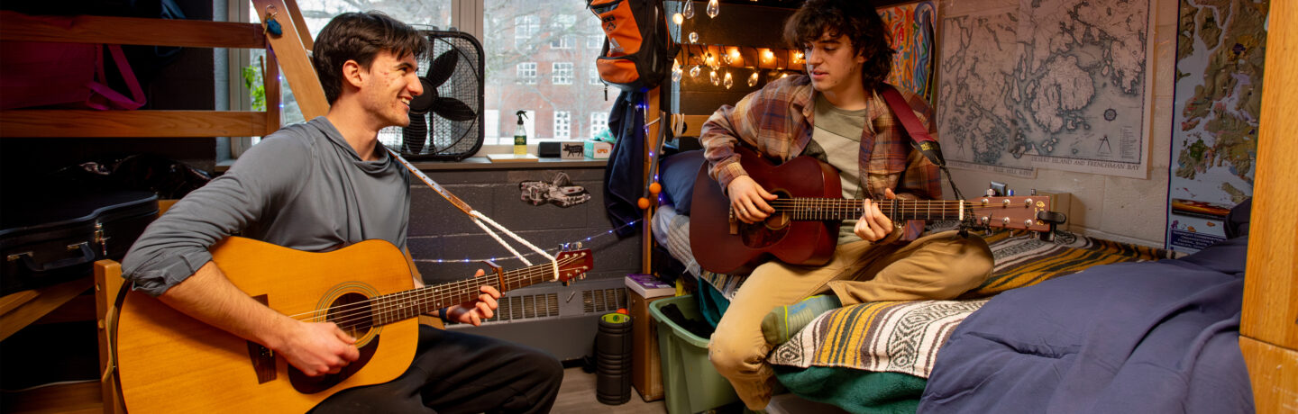 Two students play acoustic guitars together in a residence hall room. One sits on a wooden chair smiling at his roommate, who sits cross-legged on a lofted bed strumming a darker guitar. The room is decorated with string lights, maps of the Maine coast, a striped blanket, and a colorful rag rug, with a large window looking out on bare trees and a brick building.