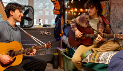 Two students play acoustic guitars together in a residence hall room. One sits on a wooden chair smiling at his roommate, who sits cross-legged on a lofted bed strumming a darker guitar. The room is decorated with string lights, maps of the Maine coast, a striped blanket, and a colorful rag rug, with a large window looking out on bare trees and a brick building.