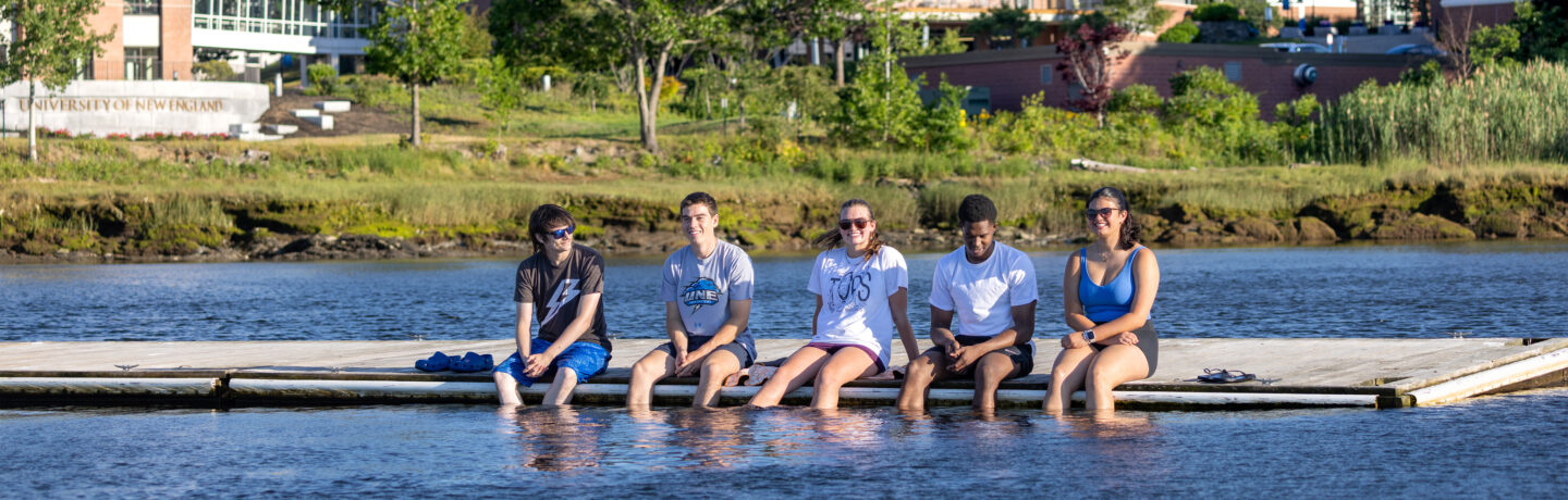 Five students sit in a row on a floating dock, smiling with their feet dangling in the river on a sunny day. Behind them, across the water, a stone sign reading University of New England stands on a grassy bank below campus buildings.