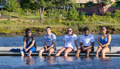 Five students sit in a row on a floating dock, smiling with their feet dangling in the river on a sunny day. Behind them, across the water, a stone sign reading University of New England stands on a grassy bank below campus buildings.