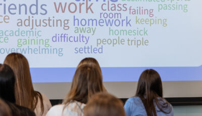 An advisor speaks to a room of students seated with their backs to the camera, gesturing toward a large projected word cloud. Words like friends, homework, adjusting, balance, homesick, independence, overwhelming, and settled appear in soft colors, reflecting students' feelings about the transition to college.