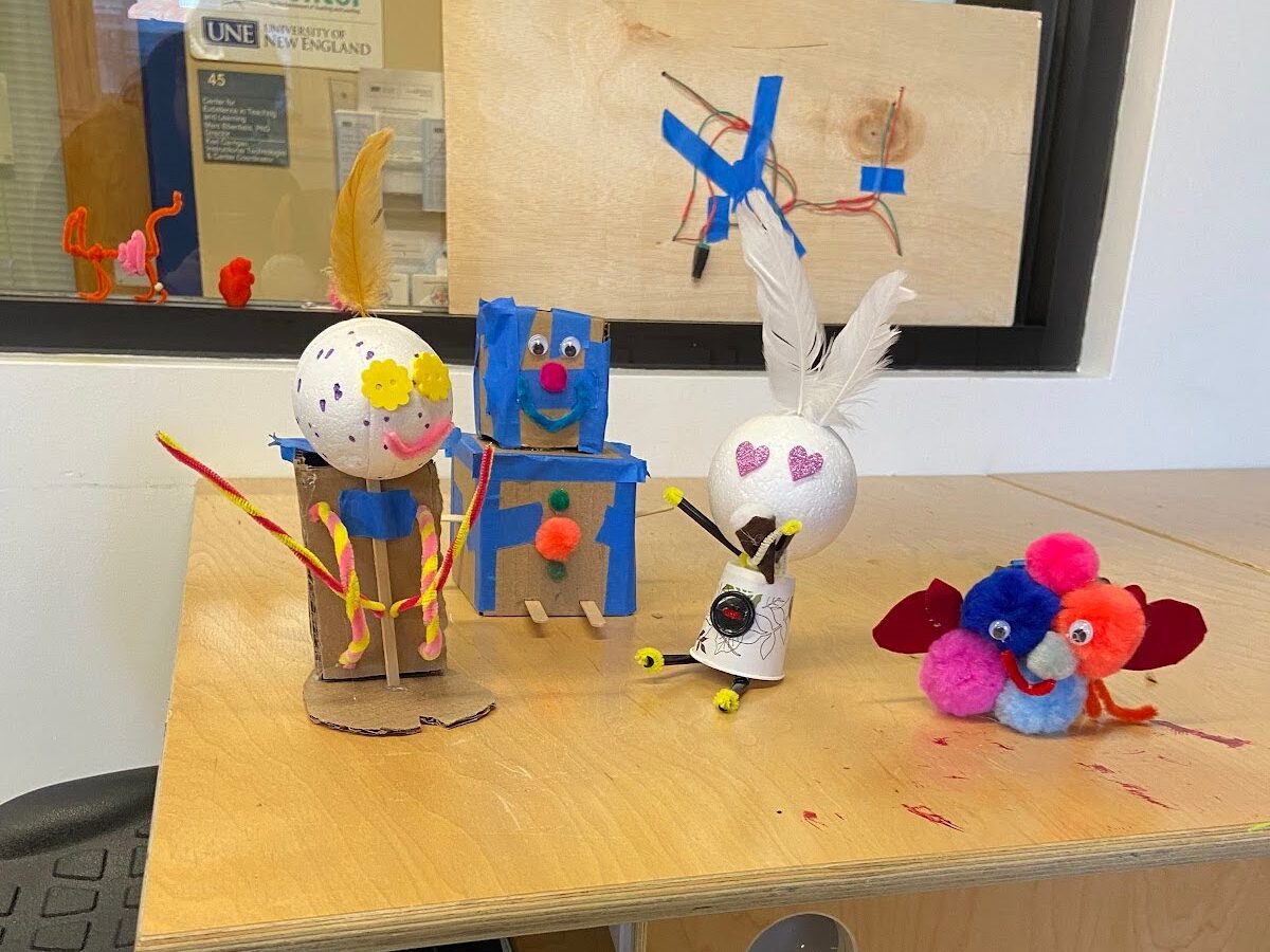 four example prototypes of "friendly robots". From left to right: a stick figure made of pipecleaners and a foam sphere wearing a backpack made out of cardboard and pipecleaners; a smiling figure made from two cardboard boxes taped together (the top box is slightly smaller to make a head); a figure made out of an upside down paper cup, pipecleaners, a foam sphere and two feathers, and a pile of pom poms with googly eyes wearing a pair of felt mittens.