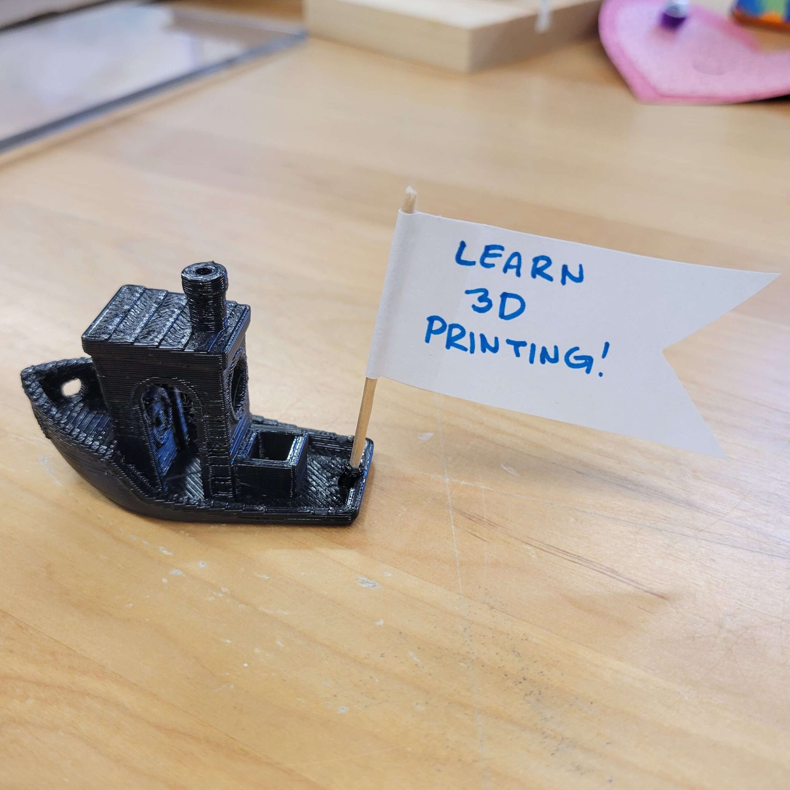 A small black 3D printed tugboat with a banner that reads "Learn 3D Printing".