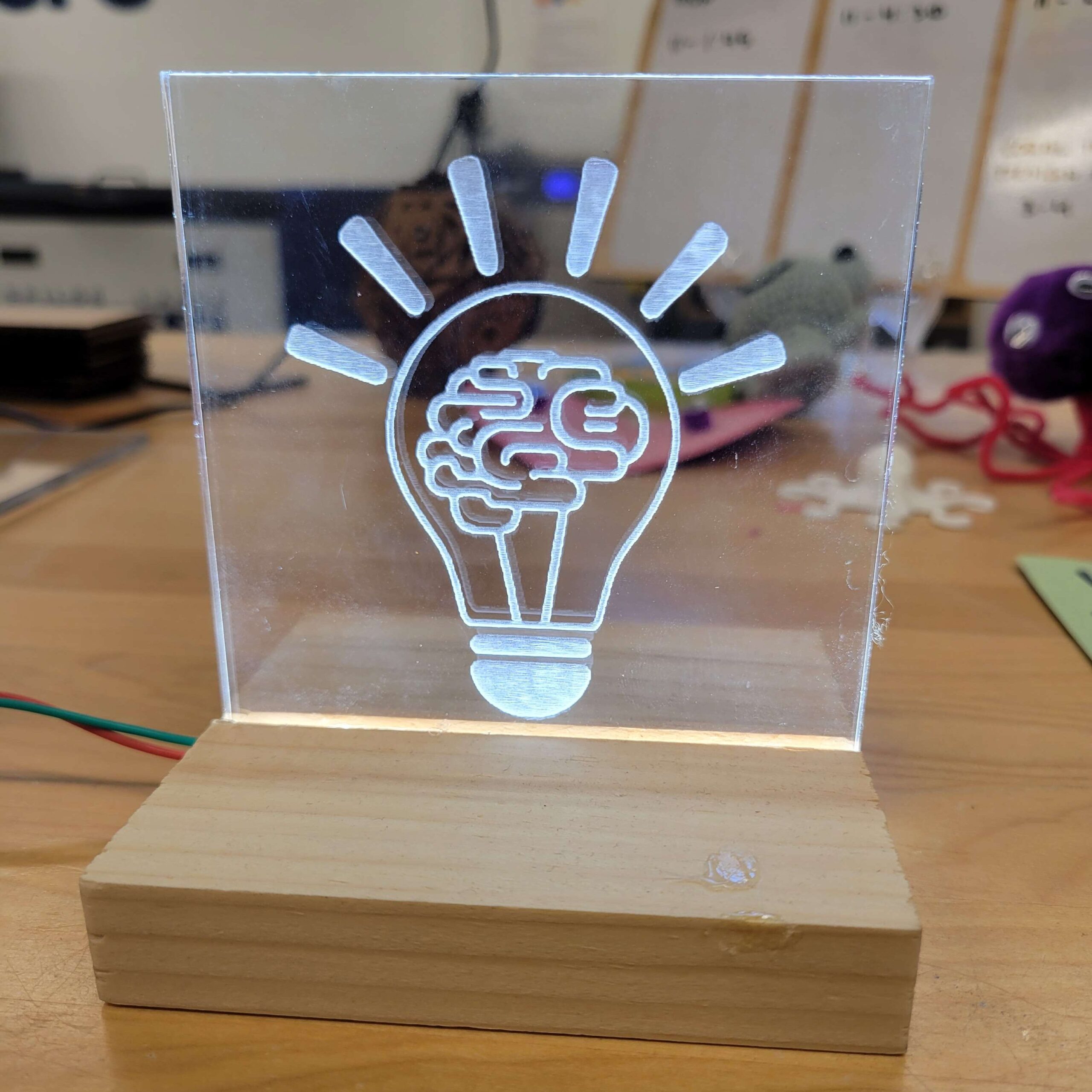 lineart of a lightbulb with a brain inside is etched into a piece of clear acrylic and is glowing.