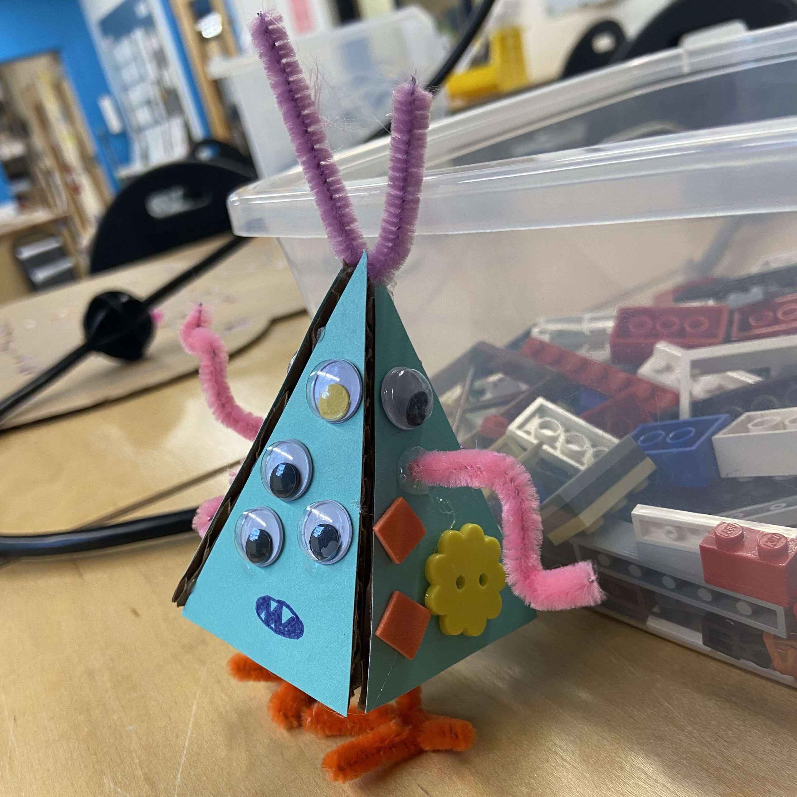 a light blue pyramid with googly eyes, multicoloured buttons, and pipe cleaner arms and legs
