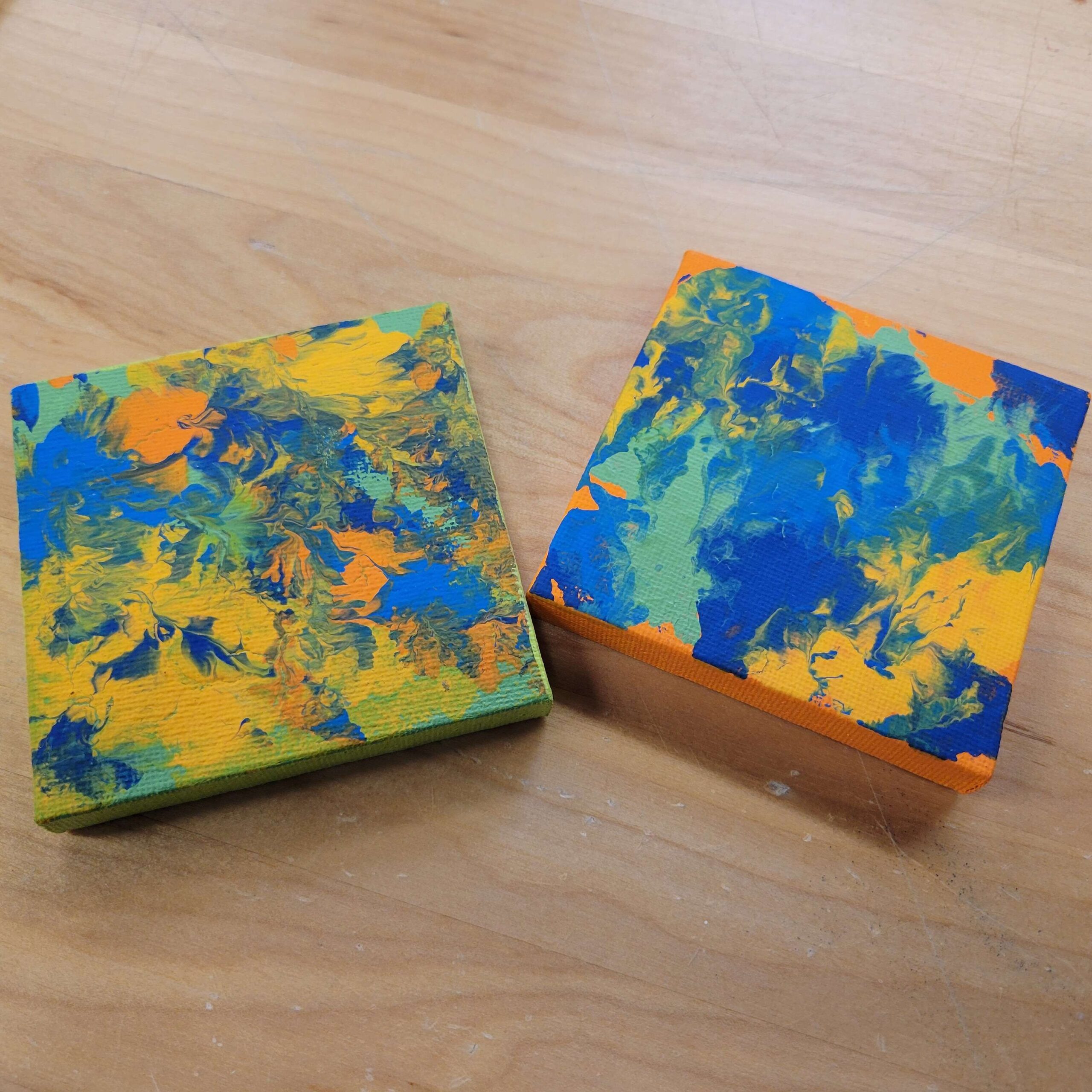 two small square canvases with blue, yellow, green, and blue paint splattered on them to make abstract patterns