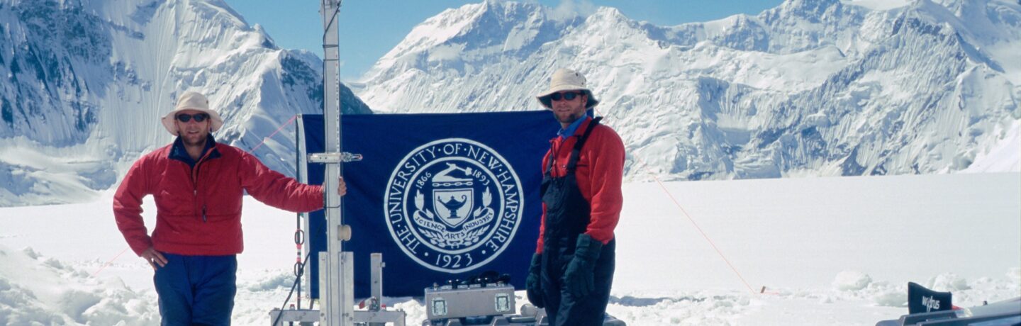 Dr. Wake and Dr. Kreutz standing at an ice core drilling site in front of a snow covered mountain